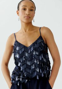 Marineblaue ärmellose Bluse mit V-Ausschnitt, die mit geschichteten, strukturierten Pailletten im Zickzackmuster verziert ist. Glatte Stoffoberfläche mit verstellbaren Trägern.