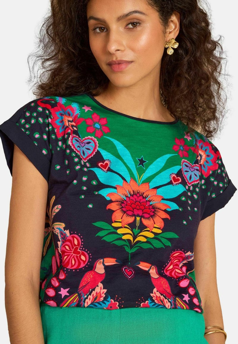 Frau trägt ein buntes T-Shirt mit Mustern aus tropischen Blumen, Herzen, Sternen und Tukan-Vögeln, kombiniert mit grünen Hosen und goldenen Blumenohrringen.
