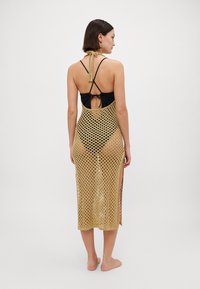 Zelta adītais pludmales pārklājs ar lielu atvērtu audumu, aizmugures saišu aizvēršanu un sānu šķēlumiem. Valkāts virs melna peldkostīma ar bez piedurknēm.