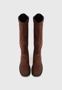 Botas de camurça castanhas até ao joelho, com uma textura suave, bico arredondado e design minimalista. Sola de borracha preta e aberturas superiores discretas.