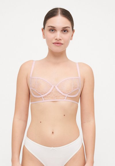 Jeune femme avec les cheveux attachés, portant un soutien-gorge brodé en rose transparent et un sous-vêtement blanc, se tenant devant un fond blanc uni.