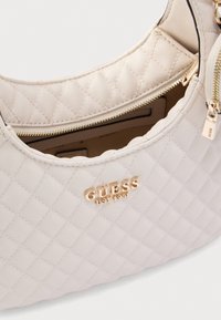 Τσάντα χειρός από κρεμ καπιτονέ ύφασμα με χρυσά μεταλλικά στοιχεία, διαθέτει κλείσιμο με φερμουάρ και ανάγλυφο λογότυπο. Μαλακή υφή με καμπυλωτό σχεδιασμό και εσωτερική τσέπη.