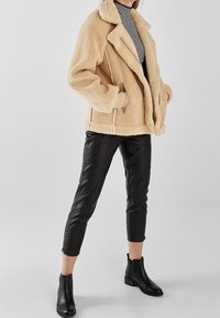 Kobieta w beżowej kurtce z owczej skóry, czarnych skórzanych spodniach 3/4 z zamkami, czarnych botkach za kostkę oraz czarno-białym swetrze z golfem w paski.