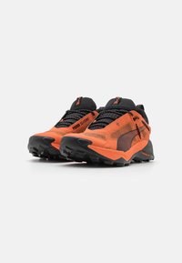 Des chaussures de sport orange avec des accents noirs, une tige en maille, une surface texturée et une semelle extérieure noire robuste avec des crampons profonds pour une meilleure adhérence.