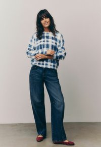 Pull en vichy bleu et blanc avec une coupe décontractée, associé à un jean large bleu marine et des ballerines rouges ornées.
