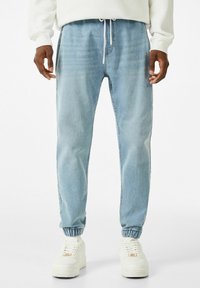 Light blue denim joggers with elastic cuffs and drawstring waistband, worn with white sneakers featuring gold accents. Smooth texture.
