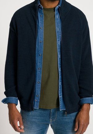 Mann trägt dunkel marineblaue Strickjacke mit Reißverschluss über blauem Denimhemd und olivgrünem T-Shirt mit blauer Jeans, Hände entspannt an den Seiten.