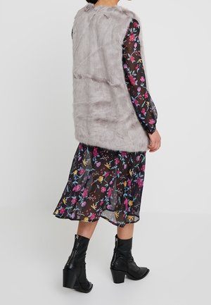 Persoon die een mouwloos grijs nepbontvest draagt over een zwarte bloemenlange mouwen jurk, gecombineerd met zwarte enkellaarsjes met hak, gezien van achteren.