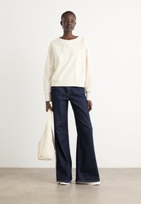 Felpa color crema con yoke testurizzato, abbinata a jeans a gamba larga blu scuro. Il modello tiene una borsa a tracolla bianca. Pavimentazione piatta e liscia.