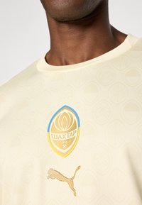 Kreminės spalvos sportinė maikė su tekstūruotu raštu, papuošta auksiniu klubo logotipu ir auksiniu Puma logotipu krūtinėje.
