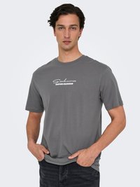 Junger Mann mit kurzen dunklen Haaren, der ein graues T-Shirt mit der Aufschrift "Sakura Blossom" trägt und mit den Händen in den Taschen einer schwarzen Jeans steht.