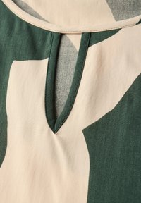 Zelená a béžová látka s vzorem, se výstřihem ve tvaru V a švovým detailem. Hladká textura zdůrazňuje barevný kontrast.