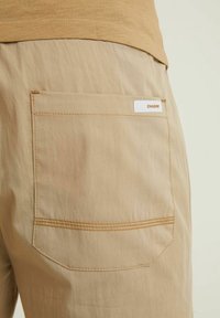 Pantalones cortos de tela color beige con un bolsillo trasero que lleva una etiqueta blanca "CHASIN'." Las costuras visibles añaden textura; material suave y ligero.