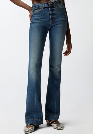 Žena ve vysokým pasem modrých zúžených zvonových džínách s odpovídajícím páskem a špičatými balerínkami se vzorem hadí kůže na jednobarevném pozadí.