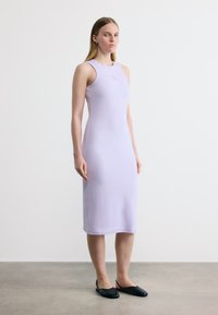 Γυναίκα που στέκεται μέσα σε κλειστό χώρο, φοράει ένα αμάνικο μεσαίο φόρεμα σε ανοιχτό μωβ χρώμα και μαύρα επίπεδα παπούτσια με λουράκι στο πίσω μέρος, μπροστά σε απλό λευκό φόντο.