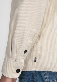 Kordhemd in Hellbeige mit einer vertikalen Rippstruktur. Verfügt über schwarze Knöpfe an den Manschetten und ein kleines Markenschild an der Seite.