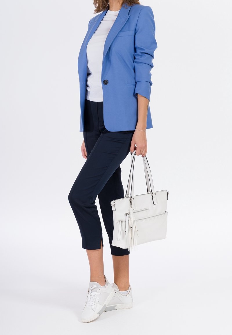 Femme portant un blazer bleu, chemise blanche, pantalon noir raccourci, baskets blanches, tenant un sac à main blanc avec détail pompons sur fond uni.
