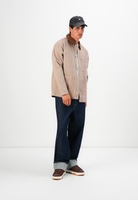 Young man wearing a beige jacket with brown collar, dark cuffed jeans, brown sneakers, and a black baseball cap, standing against white background.