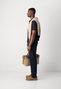 Beige strikket genser over en marineblå polo, i kombinasjon med marineblå bukser og beige sko. Holder en beige duffelbag med to hndtak.
