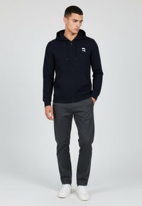 Felpa con zip blu navy con un logo sul petto, abbinata a pantaloni grigi e sneakers bianche, caratterizzata da un taglio moderno e un design casual.