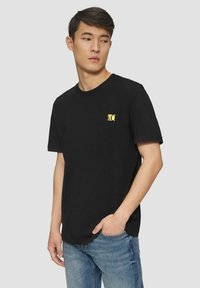 Black cotton t-shirt featuring a small yellow logo on the left chest. Short sleeves and a crew neckline. Worn with blue jeans.