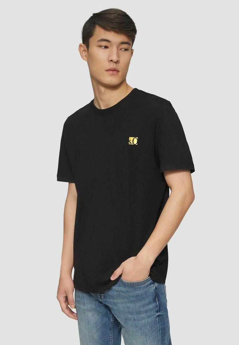 Black cotton t-shirt featuring a small yellow logo on the left chest. Short sleeves and a crew neckline. Worn with blue jeans.