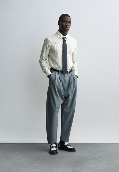 Άνδρας που φοράει κρεμ πουκάμισο με ρίγες, σκούρα γραβάτα, γκρι πλισέ παντελόνια και ασπρόμαυρα παπούτσια, στέκεται με τα χέρια στις τσέπες μπροστά σε γκρι φόντο.