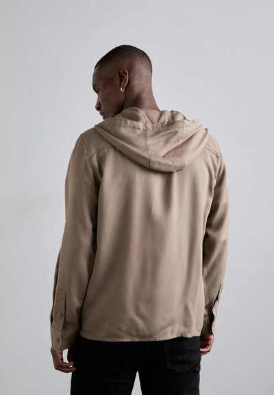 Άνδρας με κοντά μαλλιά και σκουλαρίκι, ντυμένος με μπεζ μπουφάν με κουκούλα και μαύρο παντελόνι, στέκεται με την πλάτη στραμμένη σε έναν απλό ανοιχτό φόντο.