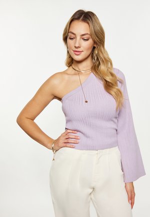 Femeie cu părul lung și blond poartă un pulover liliac cu o singură umăr și pantaloni albi cu talie înaltă, pozând cu o mână pe șold.