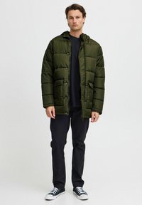 Olivenfarget polstret jakke med glidelås og trykknapper, med hette, frontlommer og kviltet design. Båret over en svart skjorte.