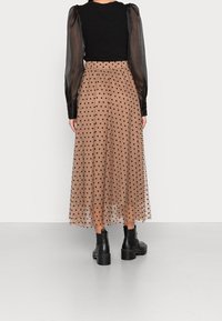 Jupe longue et transparente marron à pois noirs, associée à un haut noir côtelé avec des manches transparentes bouffantes, et des bottines noires.