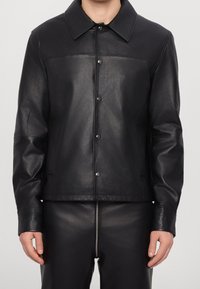 Black leather jacket featuring a pointed collar, snap buttons, long sleeves, and a smooth texture. Worn with matching pants.