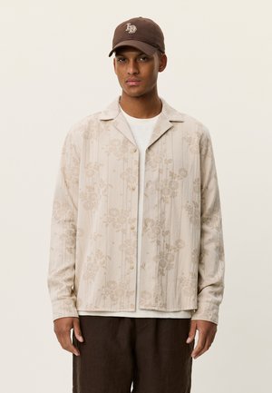 Beige langermet skjorte med blomsterprint, krage og knapper, brukt over en hvit t-skjorte, kombinert med en brun cap og mørke bukser.