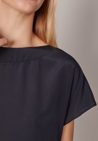 Marinblus med en mjuk, silkeslen yta. Har en rund halsringning och korta, lösa ärmar. Subtil drapering vid axlarna.