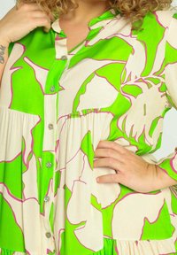 Groene en crèmekleurige blouse met knopen en een opvallend bloemmotief, met roze contouren en geplooide details. Zachte stof en een ontspannen pasvorm.