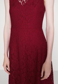 Burgundyröd spetsklänning med blommönster, ärmlös design, texturerad tyg och åtsittande midja, som visar upp intrikata detaljer och hantverk.