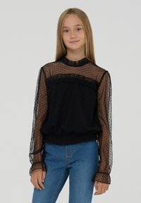 Blusa nera in tessuto trasparente a pois, con scollatura arricciata e vita elastica. Le maniche lunghe presentano polsini arricciati. Indossata con jeans blu.