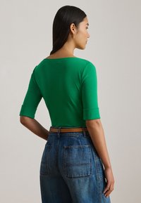 Zelený priliehavý top s tromištvrťovými rukávmi, zaobleným výstrihom a hladkou textúrou, v kombinácii s modrými džínsami s vysokým pásom a širokými nohavicami.