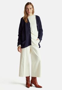 Námořnický pletený cardigan s uvolněným střihem, třemi knoflíky a kapsami, v kombinaci se krémovým svetrem a širokými bílými kalhotami, doplněný hnědými kotníkovými botami.