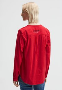 Rotes Langarmhemd aus leichtem Stoff mit einem Rückenpasse und Falten-Detail. Mit einem bedruckten Textdesign in der Nähe des Ausschnitts.