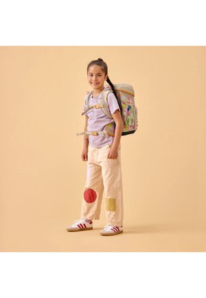 Young girl standing and smiling, wearing a colorful backpack, light purple shirt, beige pants with patches, and white sneakers with red stripes.