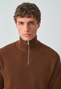 Suéter marrón de punto acanalado con cuello alto y una cremallera plateada. La tela tiene una textura suave y una silueta ajustada.