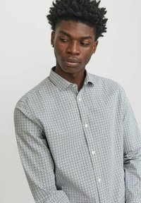 Camicia button-up con una base bianca, caratterizzata da un motivo geometrico blu e giallo. Maniche lunghe e colletto standard. Tessuto con una texture liscia.
