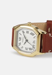 Montre Fossil avec un boîtier octagonal en or, un cadran blanc, des chiffres romains, des détails gris et un bracelet en cuir brun avec des coutures blanches.