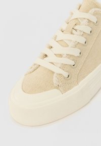 Sneaker in tela beige con lacci bianchi, tessuto texturizzato, punta arrotondata e suola in gomma con design rigato e occhielli in argento.