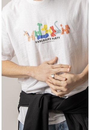 T-shirt blanc avec des illustrations colorées d'animaux en ballon au-dessus du texte "EXPLOSIVE PARTY KAOTIKO SOCIETY", mains jointes à la taille.