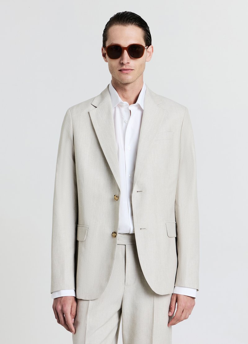 Blazer beige clair avec deux boutons, revers crantés et poches latérales. Associé à une chemise blanche et des lunettes de soleil marron, se tenant contre un fond uni.