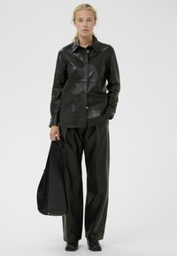 Črna usnjena srajca s koničastim ovratnikom in sprednjimi gumbi, v kombinaciji z ohlapnimi črnimi usnjenimi hlačami in usklajenim nahrbtnikom.