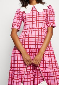 Femme portant une robe à carreaux rose et rouge avec des manches bouffantes et un col en dentelle blanc, tenant un petit sac à main blanc.