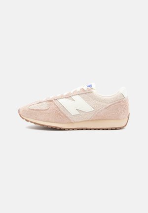 Beige og lyserød tekstureret sneaker med hvidt "N" logo, snøredesign og gummisål, vist i sideprofil på en ensfarvet baggrund.